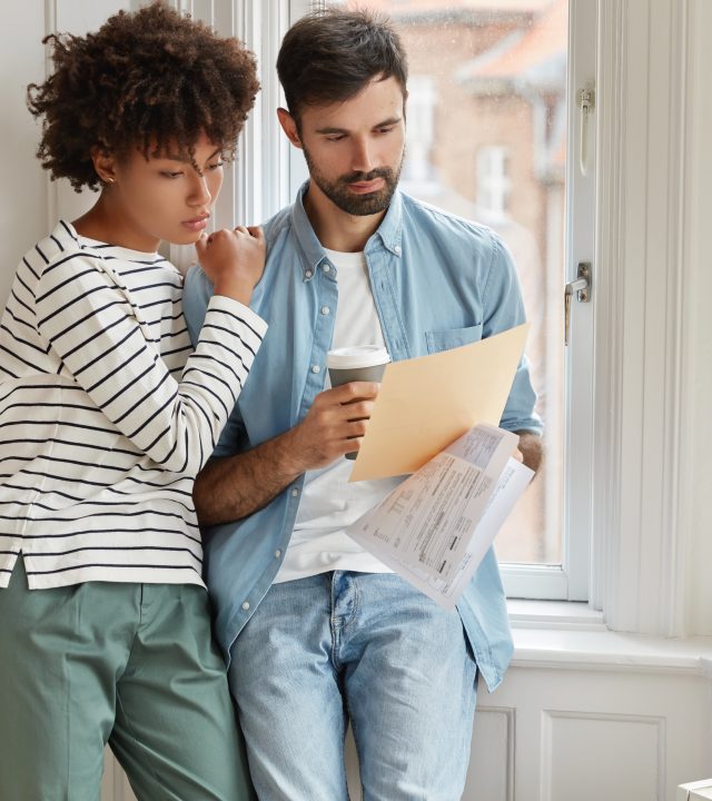 interracial young woman and man make accounting of their expenses for year, concentrated on paper work, drink takeaway coffee, pose near window, enjoy domestic atmosphere, drink hot beverage